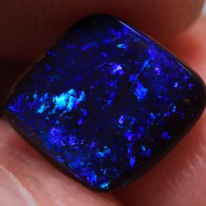 Könnte beinhalten: Ein quadratisch geschliffener schwarzer Opal-Edelstein mit lebendigen blauen und türkisfarbenen Lichtblitzen. Die polierte Oberfläche reflektiert das Licht und zeigt ein Farbspiel. Der Edelstein wird zwischen Fingern gehalten, was seine Größe und Details hervorhebt.