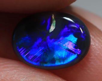 May include: A polished, oval-shaped black opal gemstone. The opal displays vibrant internal colors, primarily blues and purples, with flashes of pink and green. The stone is held between fingers, showcasing its iridescent play-of-color.