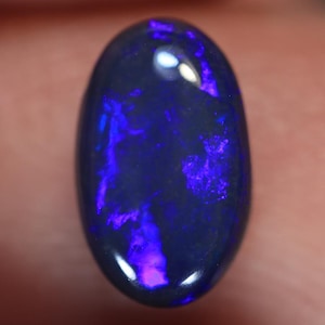 May include: An oval-shaped black opal gemstone featuring a smooth, polished surface. The stone displays vibrant, iridescent flashes of deep blue and bright purple light across its dark, opaque body, creating a distinct play-of-color effect.