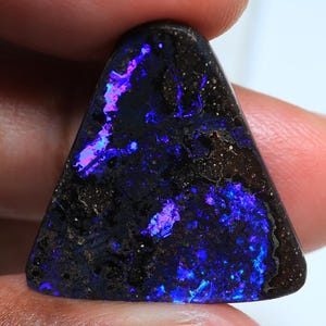 Könnte beinhalten: Ein polierter, dreieckiger Edelstein mit dunkler Grundfarbe. Der Stein weist lebhafte blaue und violette Blitze auf, die einen auffälligen Kontrast erzeugen. Der Edelstein wird zwischen den Fingern gehalten und zeigt sein einzigartiges Farbspiel und seine Textur.