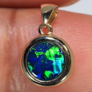 May include: A gold pendant with a round, iridescent opal stone. The opal displays vibrant green and blue colors. The pendant has a simple, elegant design, with a loop for a chain.
