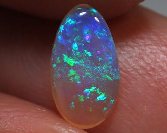 May include: A polished, oval-shaped opal gemstone. The opal displays a vibrant play-of-color, with flashes of blue, green, and purple. The stone has a translucent appearance and is held in a hand.
