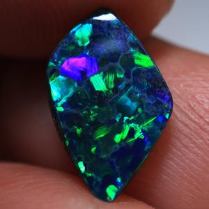 Könnte beinhalten: Ein polierter, marquiseförmiger schwarzer Opal-Edelstein mit dunkler Grundfarbe und lebhaften, irisierenden Blitzen in Neongrün, Elektroblau und hellem Violett. Die Oberfläche zeigt ein ausgeprägtes Wabenmuster im Farbspiel.