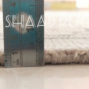 May include: Close-up of the edge of a white rug with a ruler measuring the pile height. The ruler shows a measurement of 1 mm. The text "SHAAD RUGS" is visible on the ruler.