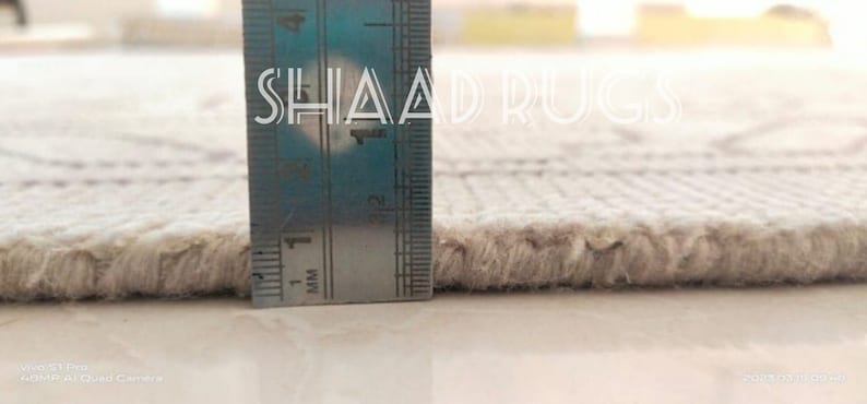 May include: Close-up of the edge of a white rug with a ruler measuring the pile height. The ruler shows a measurement of 1 mm. The text "SHAAD RUGS" is visible on the ruler.