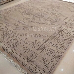 May include: A beige rug with a brown geometric pattern. The rug has a fringe border and is made of wool. The rug is called "Shaad Rugs".
