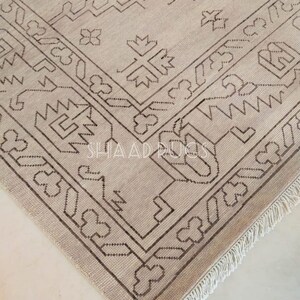 May include: A beige rug with a brown geometric pattern. The rug has a fringe border.
