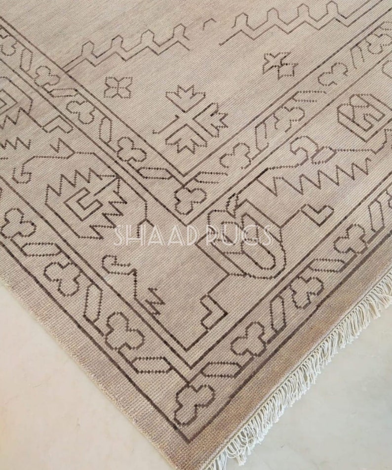 May include: A beige rug with a brown geometric pattern. The rug has a fringe border.