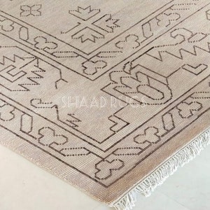 May include: A beige rug with a brown geometric pattern and fringe. The rug has a traditional design with intricate details.