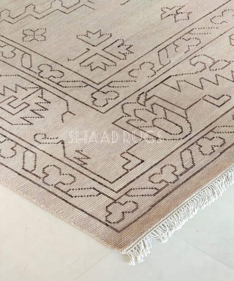 May include: A beige rug with a brown geometric pattern and fringe. The rug has a traditional design with intricate details.
