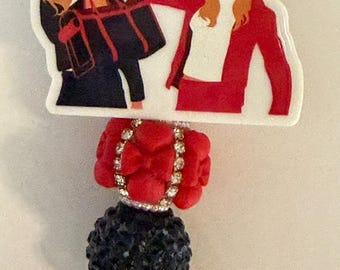 Puede incluir: Un portatarjetas con una pieza superior de acrílico blanco con el texto HOLD MY POODLE y una ilustración de dos figuras. Debajo hay cuentas texturizadas rojas y negras, un pequeño dije de flor negra y un clip circular.