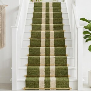 May include: A green and cream striped stair runner on a white staircase. The runner has brass rods and is flanked by white banisters. A macrame wall hanging and potted plants are also visible.