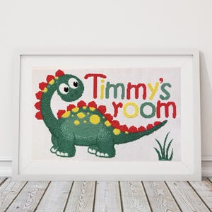 May include: Framed cross-stitch artwork depicting a green dinosaur with red and yellow details. The words "Timmy's room" are stitched in red, yellow, and green. The artwork is presented in a white frame, suitable for a child's bedroom.