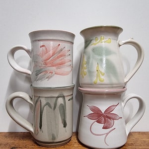 May include: Four ceramic mugs with different floral designs. The mugs are stacked on top of each other. The top mug has a yellow flower design, the second mug has a pink flower design, the third mug has a green and white abstract design, and the bottom mug has a red flower design.