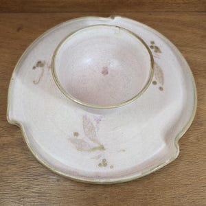 May include: A handmade ceramic serving dish set. The set includes a shallow, round plate with a small, centered bowl. The plate has a light pink glaze with subtle floral accents and a small notch on the side. The bowl is also light pink.