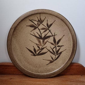 May include: A round ceramic plate with a brown glaze and a black design of stylised bamboo leaves.