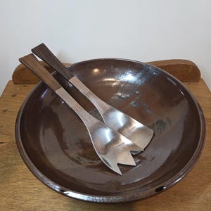 May include: A large, round, dark brown glazed ceramic bowl containing two salad servers. The servers feature polished silver-toned metal heads with three-pronged fork ends and straight, dark brown wooden handles that extend from the metal base.