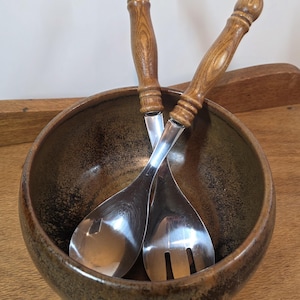 May include: A round, dark brown ceramic bowl with a speckled finish containing a pair of salad servers. The servers feature polished stainless steel heads and rounded, light brown wooden handles with a visible wood grain pattern.