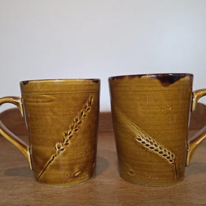 May include: Two cylindrical ceramic mugs with a glossy mustard-brown glaze. Each mug features a textured, embossed wheat stalk design on the front surface. The mugs have a smooth finish, a sturdy handle on the side, and a slightly darker brown rim.