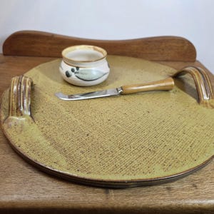May include: A handmade ceramic serving tray with a textured, yellow-green surface. It features two curved handles, a small ceramic bowl with a floral design, and a cheese knife with a wooden handle. The tray is on a wooden surface.