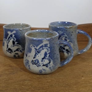 May include: Three ceramic mugs with a blue and white floral design. The mugs are different sizes and have handles. The mugs are sitting on a wooden surface.