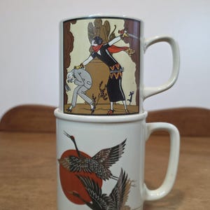 May include: Two stacked ceramic mugs with off-white bases. The top mug features an illustration of a person in a black dress holding a sword over a person in a grey suit. The bottom mug displays two black cranes flying in front of a large red circle.