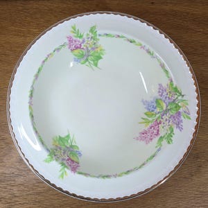 May include: A circular, cream-coloured ceramic plate with a floral design. The rim has a gold trim and a scalloped edge. The floral pattern features purple, lavender, yellow, and green flowers arranged around the plate's edge.