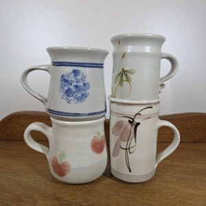 May include: Four ceramic mugs stacked on a wooden surface. The mugs are off-white with hand-painted designs: blue floral patterns, green and yellow sprigs, red apples, and abstract pink and black floral motifs. Each mug has a curved handle.