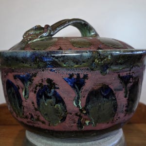 May include: A round ceramic lidded bowl featuring a matte mauve base with a repeating pattern of dark blue and olive green glazed floral motifs. The lid includes a curved handle with matching blue and green glaze drips along the rim.