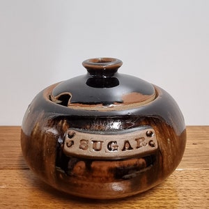 May include: A brown and black ceramic sugar bowl with a lid. The bowl has a label that says "SUGAR".