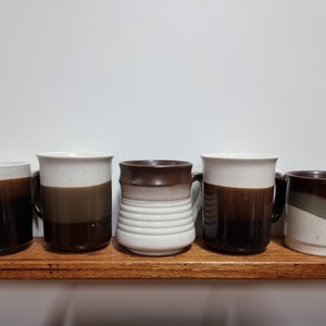May include: Five ceramic coffee mugs with brown and white glaze. The mugs have different designs and are arranged in a row on a wooden shelf.