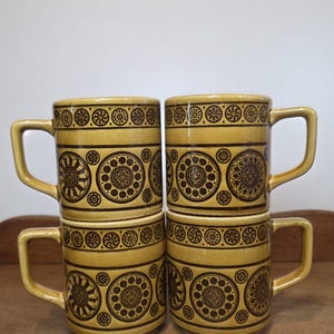 May include: A set of four cylindrical ceramic mugs with a mustard yellow glaze and dark brown printed geometric patterns. Each mug features a repeating circular motif around the centre and a smaller dotted band near the top rim with a handle.
