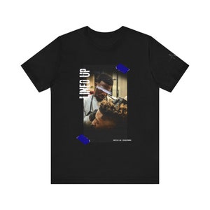 May include: Black t-shirt with a photo of a person getting a haircut. The photo is surrounded by blue tape and the text "LINED UP" is printed vertically on the left side of the photo.