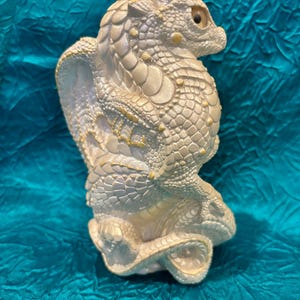 May include: A cream-colored dragon figurine with detailed scales and a textured surface. The dragon is in a seated position with its head turned to the side. The figurine has yellow accents and is set against a teal background.