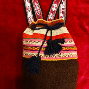 May include: A woven backpack with a drawstring closure and shoulder straps. The bag features a brown base, a striped central section with pink, orange, and white, and decorative straps with geometric patterns in red, white, and black. Dark blue tassels hang from the drawstring.