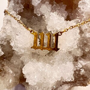 May include: Gold chain necklace with a large, ornate, gold letter 'W' pendant. The necklace is displayed on a white, crystal-like surface.