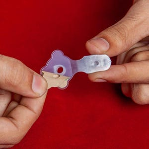 Pode incluir: Um objeto transparente em forma de chave de plástico com uma cabeça em forma de urso roxo e uma cauda retangular. O objeto tem um buraco na cabeça do urso e um pequeno pedaço de papel castanho preso na parte de trás. O fundo é vermelho liso.