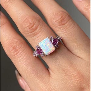 May include: A silver ring with a large, rectangular opal centre stone. The opal has a milky white appearance with iridescent flashes. Smaller pink gemstones flank the opal, adding colour. The ring is set on a silver band.