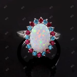 May include: A silver ring with an oval opal, displaying iridescent colours, encircled by alternating red and blue gemstones. The ring is set against a dark background, accentuating the vibrant colours of the stones.