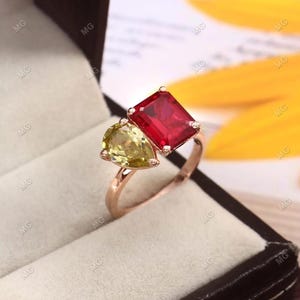 Könnte beinhalten: Roségoldener Ring mit einem birnenförmigen gelben Edelstein und einem rechteckigen roten Edelstein. Der Ring ist in einem zarten Band gefasst und wird in einer Schmuckschatulle präsentiert.
