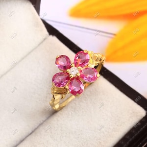 Op de afbeelding: Een goudkleurige ring met een bloemdesign met vijf roze edelstenen en een kleine, heldere edelsteen in het midden. De ring is gezet in een bloempatroon en de band heeft een textuur.