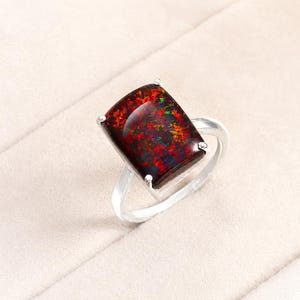 Op de afbeelding: Een zilveren ring met een rechthoekige zwarte opaal edelsteen met levendige rode, groene en blauwe iriserende flitsen. De steen is vastgezet met vier metalen pootjes op een gepolijste zilveren band, met een glad en glanzend oppervlak.