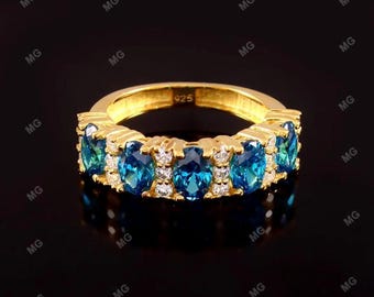 Puede incluir: Un anillo dorado con una serie de piedras preciosas azules ovaladas y piedras blancas más pequeñas. El anillo está engastado sobre un fondo oscuro, resaltando el brillo de las piedras. El anillo está marcado con "925".