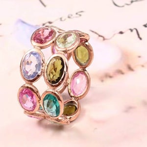 Puede incluir: Un anillo de oro rosa con una doble fila de piedras preciosas ovaladas. Las piedras preciosas son de varios colores, incluyendo rosa, verde, azul y amarillo.