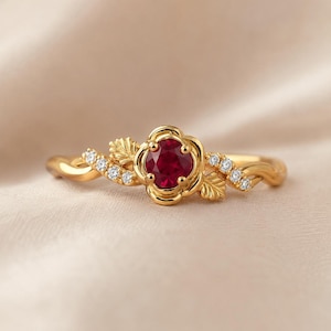 Könnte beinhalten: Ein goldener Ring mit einem zentralen, rund geschliffenen, tiefroten Rubin in einer blütenförmigen Fassung. Die Ringschiene ist beidseitig mit kleinen klaren Steinen und skulpturalen Blattdetails verziert, was ein poliertes Design ergibt.