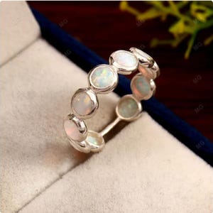May include: A silver band ring featuring a series of round, bezel-set white opals that display iridescent flashes of color. The stones are arranged in a continuous row around the circumference of the metal band, creating a circular design.