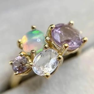 May include: A gold ring featuring an opal, two amethyst, and a clear gemstone. The opal has a pearlescent sheen, while the amethysts are a deep purple. The clear gemstone is oval-shaped. The ring's setting is gold.