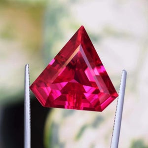 Op de afbeelding: Een helderrode, driehoekige edelsteen, vastgehouden door een pincet. De facetten van de edelsteen vangen het licht op, waardoor een sprankelend effect ontstaat. De achtergrond is onscherp, met groene en gele tinten.