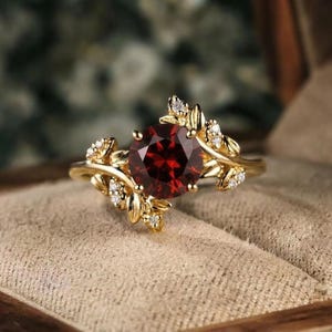 Könnte beinhalten: Ein goldener Ring mit einem großen, runden, tiefroten Granat-Edelstein in der Mitte. Das Band ist mit filigranen goldenen Blattmotiven gestaltet, die vom Zentrum ausgehen und mit kleinen, klaren, rund geschliffenen weißen Steinen besetzt sind.