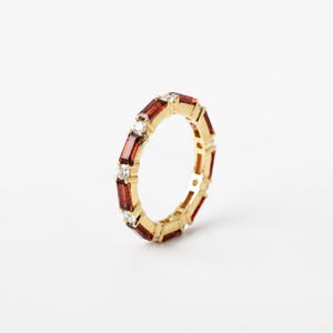 Könnte beinhalten: Ein Gold-Eternity-Ring mit einem abwechselnden Muster aus rechteckigen, im Baguetteschliff geschliffenen, tiefroten Granat-Edelsteinen und kleinen, runden, im Brillantschliff geschliffenen, klaren weißen Diamanten in einer Ringschiene aus poliertem Gelbgold.